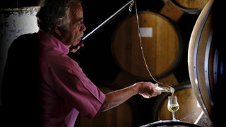 A man in a pink shirt pours wine from a bottle into a glass in a dimly lit wine cellar with wooden barrels in the background.