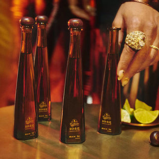 Four sleek Don Julio 1942 Añejo Tequila 50ml bottles sit on a table. A jeweled hand reaches for them, while lime wedges and golden lighting create an inviting scene. Notes of caramel and toasted oak fill the air.