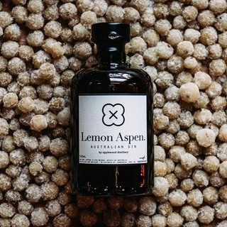 A dark glass bottle of Applewood Lemon Aspen Gin 500ml sits on a surface sprinkled with round, sugar-coated lemon aspen berries. Its white label has black text and a minimal flower-like logo. This gin features citrus botanicals.