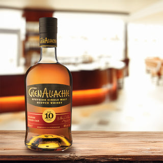 A bottle of GlenAllachie 10 Year Old Spanish Virgin Oak Single Malt Scotch Whisky rests on a wooden surface, with a modern, blurred bar background, hinting at aromas of orchard fruits and baking spices.
