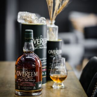 A bottle of Overeem Port Cask Matured – Cask Strength Tasmanian Single Malt Whisky sits on a wooden table beside its branded glass and cylindrical packaging, with dried flowers nearby and aromas of butterscotch and spiced dried fruit in the air.