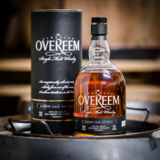 A bottle of Overeem Sherry Cask Matured – Distiller’s Strength Tasmanian Single Malt Whisky, with its cylindrical packaging, sits on a round metal tray against a blurred wooden background—evoking caramel and dark chocolate notes.
