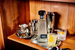 Bartending setup with 1800 tequila bottle, cocktail shaker, and glass with lemon wedge on a wooden shelf.