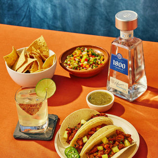 A table set with two tacos, tortilla chips, salsa, green sauce, a citrus cocktail with lime, and a bottle of 1800 Blanco Tequila 700ml made from fine agave rests atop an orange tablecloth.