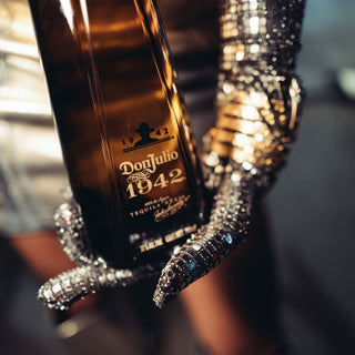 A close-up of a hand in a sparkling silver sequined glove holding a Don Julio 1942 Añejo Tequila 1.75L bottle, its label visible amid notes of roasted agave and oak sweetness.