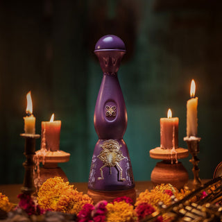 The Clase Azul Dia de Los Muertos 2024 Música Limited Edition Tequila 1L, in a tall deep purple bottle adorned with gold oak-inspired designs, is displayed with marigold flowers and candlelit wooden holders for a festive ambiance.