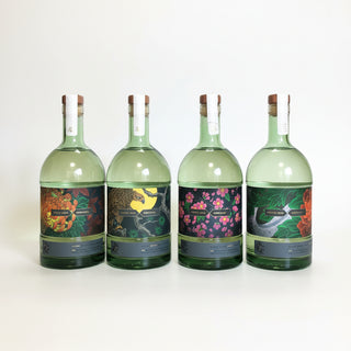 Four green glass bottles of Japanese sake lined up on a white surface, featuring colorful traditional labels with floral and bird designs, set against a dark background with draped white fabric.