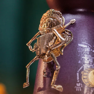 A metallic skeleton figurine with a hat plays an accordion and holds a guitar, adorning the Clase Azul Dia de Los Muertos 2024 Música Limited Edition Tequila 1L bottle, set against a blurred green background and purple patterned surface.