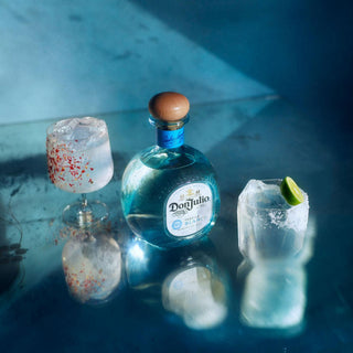 A 50ml bottle of Don Julio Blanco Tequila stands on a reflective blue surface between two cocktails—one in a tall glass garnished with red pepper flakes, the other in a lowball glass with a salted rim and citrus zest.