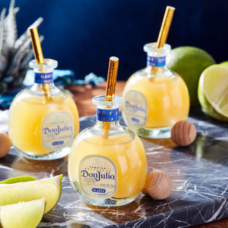 Three 50ml Don Julio Blanco Tequila bottles are filled with a yellow cocktail, featuring sweet agave and citrus zest notes. Each has a gold straw and is set on marble with lime wedges and wooden stoppers nearby.