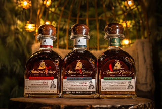 Three bottles of Admiral Rodney rum syrup arranged side by side on a rustic wooden surface, with a warm, blurred tropical background.