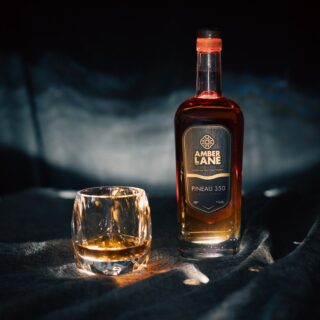 A bottle of Amber Lane Pineau 350 Australian Single Malt Whisky with a dark label stands beside a glass of amber whisky on dark fabric, spotlighted—aromas of red fruit and cinnamon linger in the air.