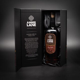 A large bottle of Amber Lane Sleigh Bells Australian Single Malt Whisky stands upright in an open black gift box with gold accents and inner text. Notes of spiced apple enrich the softly lit, dark background.