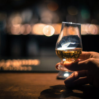 A hand holds a glass of toffee-hued Amrut Indian Single Malt Whisky on a wooden bar counter, set against a blurred background of warm, glowing lights.