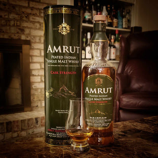 A bottle and cylindrical box of Amrut Peated Cask Strength Indian Single Malt Whisky sit on a table with a filled glass, the cozy background evoking peat smoke, fireplace warmth, shelves, and a leather chair.