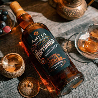 A bottle of Amrut Portonova Indian Single Malt Whisky rests on a wooden table beside ornate cups, a tea glass, and grapes, bathed in warm light that emphasizes its oaky notes and rich fruit character.