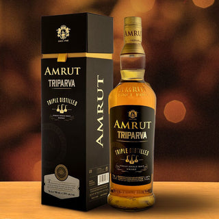 A bottle of Amrut Triparva Indian Single Malt whisky is placed beside its elegant black and gold box on a wooden surface, set against a softly blurred, warm-toned background.