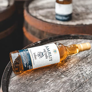 A bottle of Amrut Raj Igala Indian Single Malt Whisky rests on its side atop a wooden barrel, suggesting creamy peach and honey notes, with a blurred background featuring another barrel and bottle.