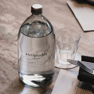 A bottle of Antipodes Sparkling Mineral Water stands next to an empty glass on a round coaster, with a folded napkin, fork, and knife arranged nearby on a marble surface.