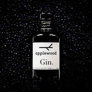 A 500ml bottle of Applewood Gin, made with native botanicals, stands upright before a dark textured background. Its minimalist white label displays black text and a simple branch graphic above the brand name.