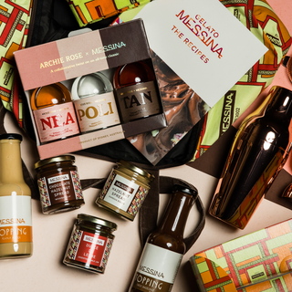 A flat lay of the Archie Rose x Messina Neapolitan Set, featuring a recipe book, cocktail shaker, chocolate gin liqueur gift set, dessert toppings, chocolate spread, and a colorful map-patterned bag.