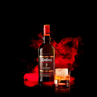 A bottle of Ardbeg 5 Year Old Wee Beastie Single Malt Scotch Whisky stands beside a whisky glass with ice, set against a black background with swirling red smoke, evoking earthy peat smoke and cracked black pepper notes.