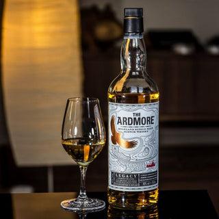 A bottle of Ardmore Legacy Single Malt Scotch Whisky sits beside a filled whisky glass on a dark surface, sweet malt aromas drifting in the softly lit background with a tall lamp.