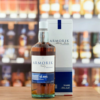 A bottle of Armorik 10 Ans (10 Year Old) French Single Malt Whisky is displayed beside its box on a wooden surface, with blurred bottle shelves behind—its flavor profile features notes of orchard fruits and honey.