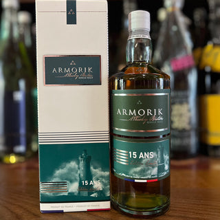 An Armorik 15 Ans French Single Malt Whisky bottle stands by its box on wood, emphasizing its subtle peat and smoky notes, with blurred bottles in the background.