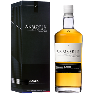 A bottle of Armorik Classic French Single Malt Whisky stands beside its black, striped box, both featuring white and gold Armorik branding and product details, hinting at fresh peach and spicy cereal aromas inside.