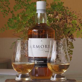 A bottle of Armorik Classic French Single Malt Whisky stands behind two filled glasses on a table, green leafy plants in the background, capturing subtle spicy cereal aromas that enhance the inviting scene.