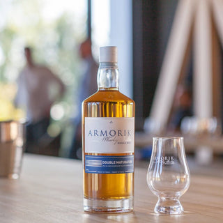 A bottle of Armorik Double Maturation French Single Malt Whisky sits beside a branded whisky glass on a wooden table, with blurred people and large windows in the background, evoking notes of vanilla spice and honey oak.