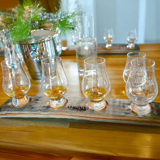 A wooden tray displays four glasses of Armorik Pineau des Charentes Single Cask French Whisky on a polished table, with a water glass, metal ice bucket with greenery, and another tray holding more glasses near dried figs and red grapes.