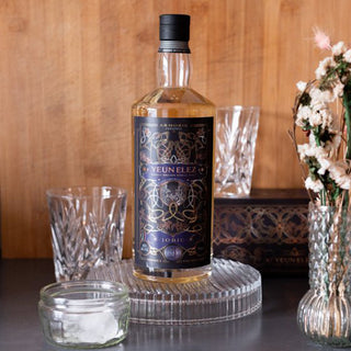 A bottle of Armorik Yeun Elez Jobic French Single Malt Whisky rests on a glass tray beside empty cut-glass tumblers, a small ice jar, and a vase of dried flowers against a wooden backdrop.