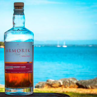 A bottle of Armorik Sherry Cask French Single Malt Whisky rests outdoors, backed by blue water, rocks, and sailboats beneath a clear sky—evoking hints of vanilla and roasted almonds.