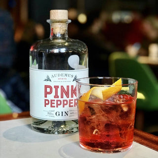 Bottle of Audemars Pink Pepper gin with a cocktail garnished with a lemon peel on a bar counter.