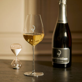 A glass of white wine, an hourglass, and a Billecart-Salmon Le Réserve NV (Gift Boxed) bottle with a simple white logo sit on a dark table. Refined bubbles and soft light evoke an elegant atmosphere.