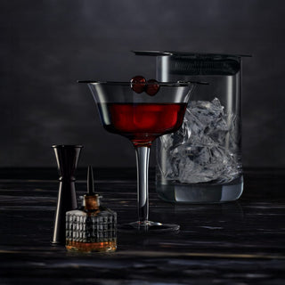 A stemmed glass with a cocktail garnished with two cherries sits beside ice-filled mixing glass, jigger, bitters bottle, and Baker’s 7 Year Old Single Barrel Bourbon—inviting notes of toasted oak and caramel.