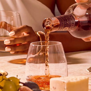 A hand pours Basil Hayden Red Wine Cask Finish Bourbon into a glass, while another holds a drink. Cheese, grapes, figs, and dried fruits are arranged on the table in a warmly lit, elegant setting.
