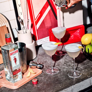 Cocktail being poured into a glass with a bottle of Beefeater gin on a bar counter.