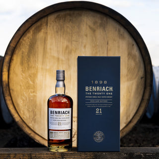 A bottle of Benriach The Twenty One Single Malt Scotch Whisky with its box is shown before a large barrel outdoors. The packaging notes summer berries, emphasizing its 21-year aging as a Speyside single malt.
