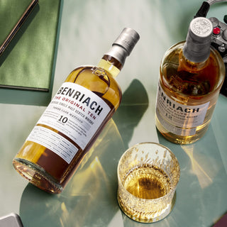 Two bottles of Benriach The Original Ten Single Malt Scotch Whisky rest on a glass table alongside a whisky glass and video game controller, inviting you to enjoy their notes of orchard fruits and gentle malt sweetness.