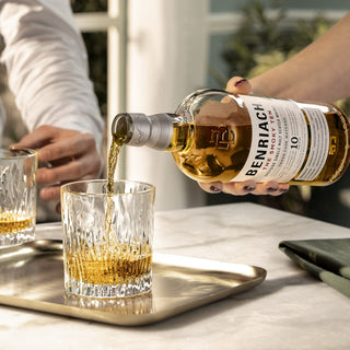 A hand pours Benriach The Smoky Ten Single Malt Scotch Whisky, known for its smoked honey notes, into a glass on a silver tray; another hand and glass rest nearby in a bright, sunlit room.