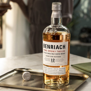A bottle of Benriach The Smoky Twelve Single Malt Scotch Whisky sits on a metal tray, its cap nearby. Aromas of smoked vanilla and dark chocolate fill the air while a blurred figure in the background holds a glass.
