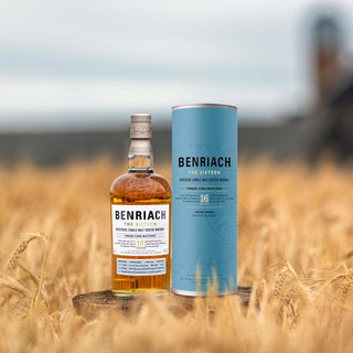 A bottle of Benriach The Sixteen Single Malt Scotch Whisky with its blue cylindrical packaging stands in a wheat field, the blurred sky evoking notes of honeyed malt and baked apples.