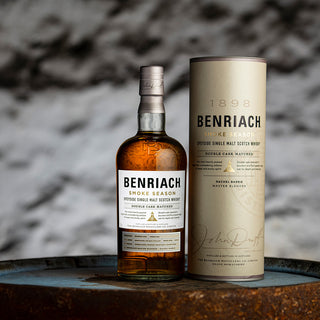 A bottle of Benriach Smoke Season Single Malt Scotch Whisky is placed next to its cylindrical box on a rusted metal surface, evoking smoky vanilla notes, with a blurred marbled gray background.