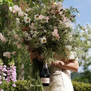 A person in a light outfit holds a lush bouquet of wildflowers that hides their face, with a bottle of Bollinger Rose NV 1.5L nestled among the blooms. Green foliage and pink flowers create a dreamy background.