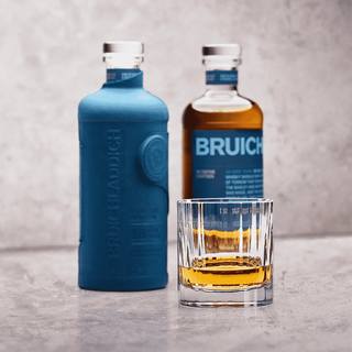 A glass of Bruichladdich 18 Year Old Single Malt Scotch Whisky sits in front of two bottles—one blue matte, one showing golden spirit—its aroma offering hints of vanilla and honeyed malt, set against a light gray textured background.