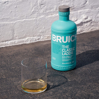 A bottle of Bruichladdich The Classic Laddie Single Malt Scotch Whisky rests on a sunlit concrete surface beside a glass, its pour highlighting creamy vanilla and honeyed sweetness.