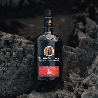 A bottle of Bunnahabhain 12 Year Old Single Malt Scotch Whisky sits on jagged dark rocks, its black and red label suggesting the nutty sweetness and subtle vanilla notes inside, set against a moody, natural backdrop.